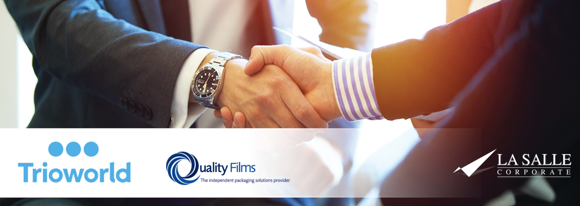 Business people shaking hands over a desk with company logos on a blue banner.