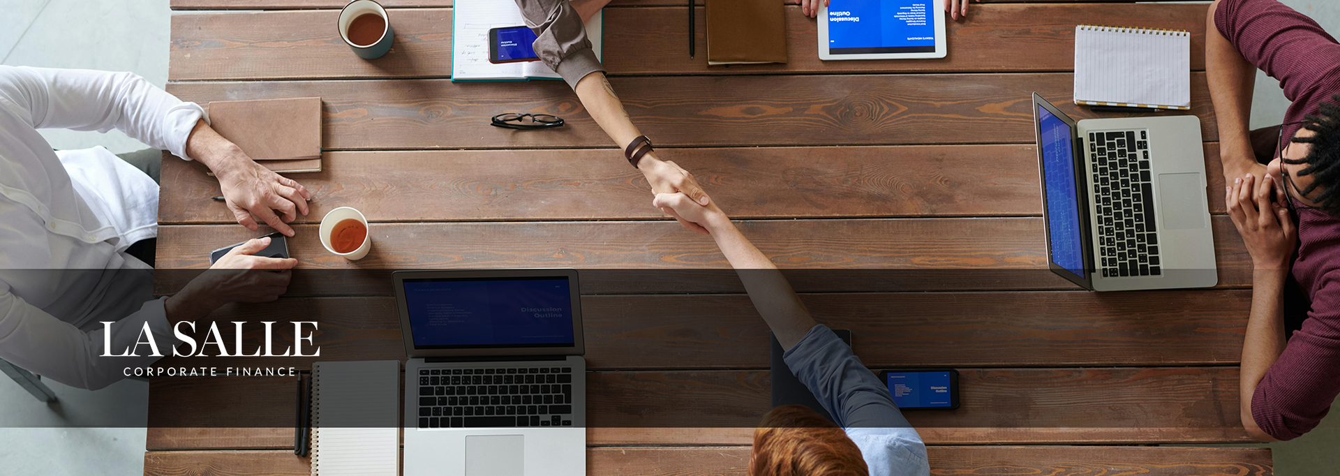 People around a wooden conference table meeting, hands reaching in for a team huddle