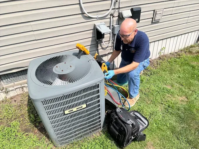 HVAC technician servicing an AC unit near a house. He's using tools on colorful hoses.