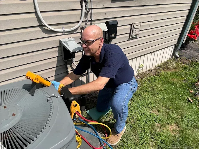 Man in blue shirt and jeans repairs an AC unit outside a home.