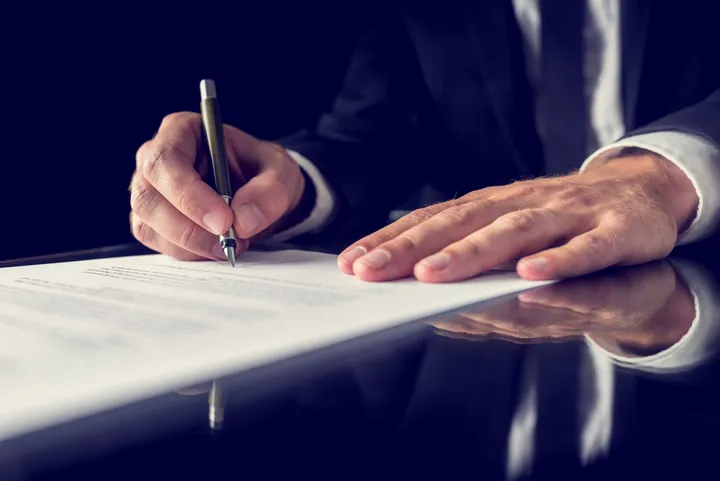 Person in suit signing a document with a pen at a dark table.