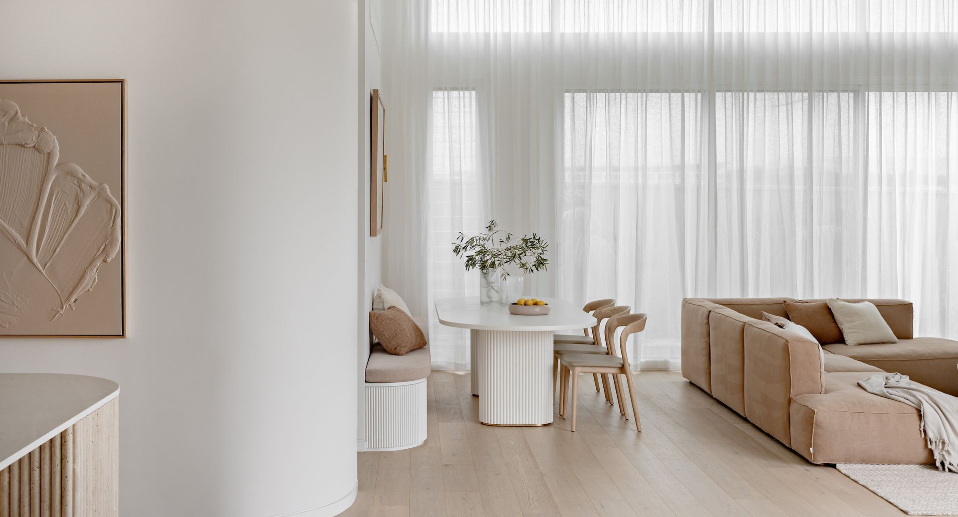 Modern, neutral-toned living space. Dining table with chairs, beige sectional sofa, and floor-to-ceiling sheer curtains — SDC Building Design In Kiama, NSW