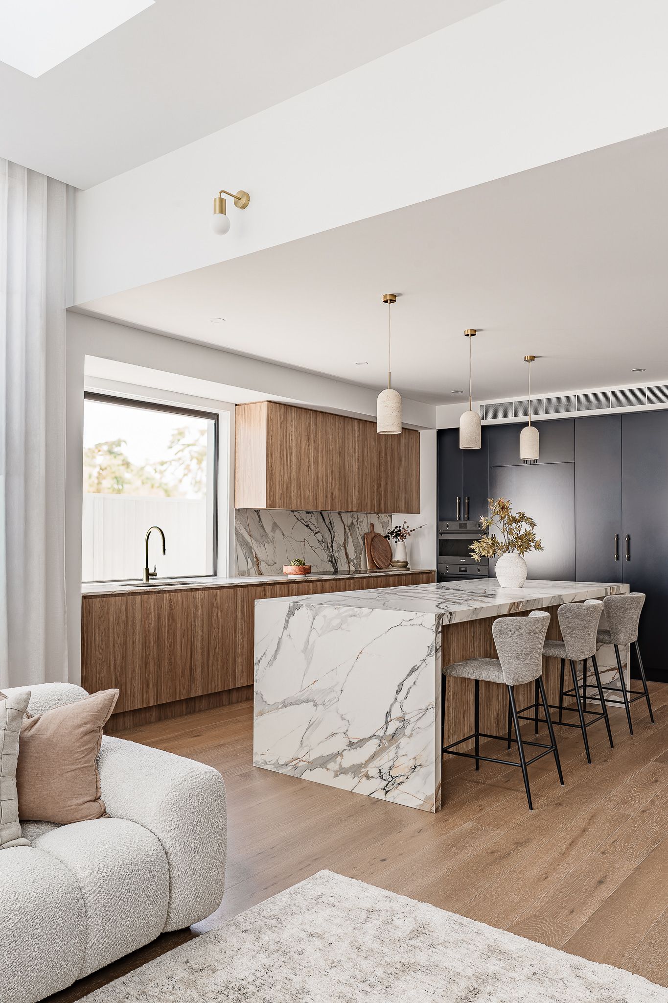 Modern kitchen with marble island, wooden cabinets, and three pendant lights; a couch in the foreground — SDC Building Design In Wollongong, NSW