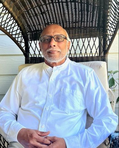 Older man with glasses in a white shirt sits on a wicker chair on a porch.