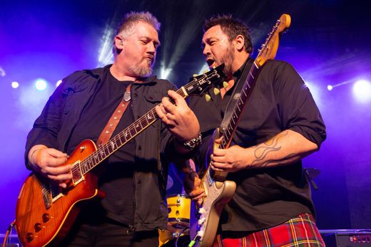 Two guitarists on stage, playing instruments intensely. One wears a kilt. Stage lit with blue.