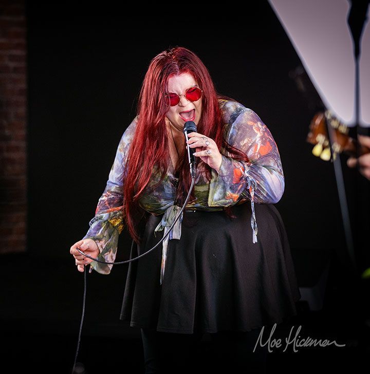 Alice Armstrong with her long red hair and red tinted glasses singing on stage