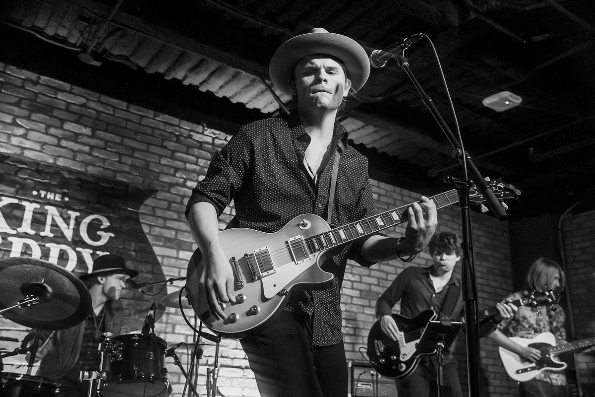 Black and white photo of Marcus Trummer playing guitar with the band in the background