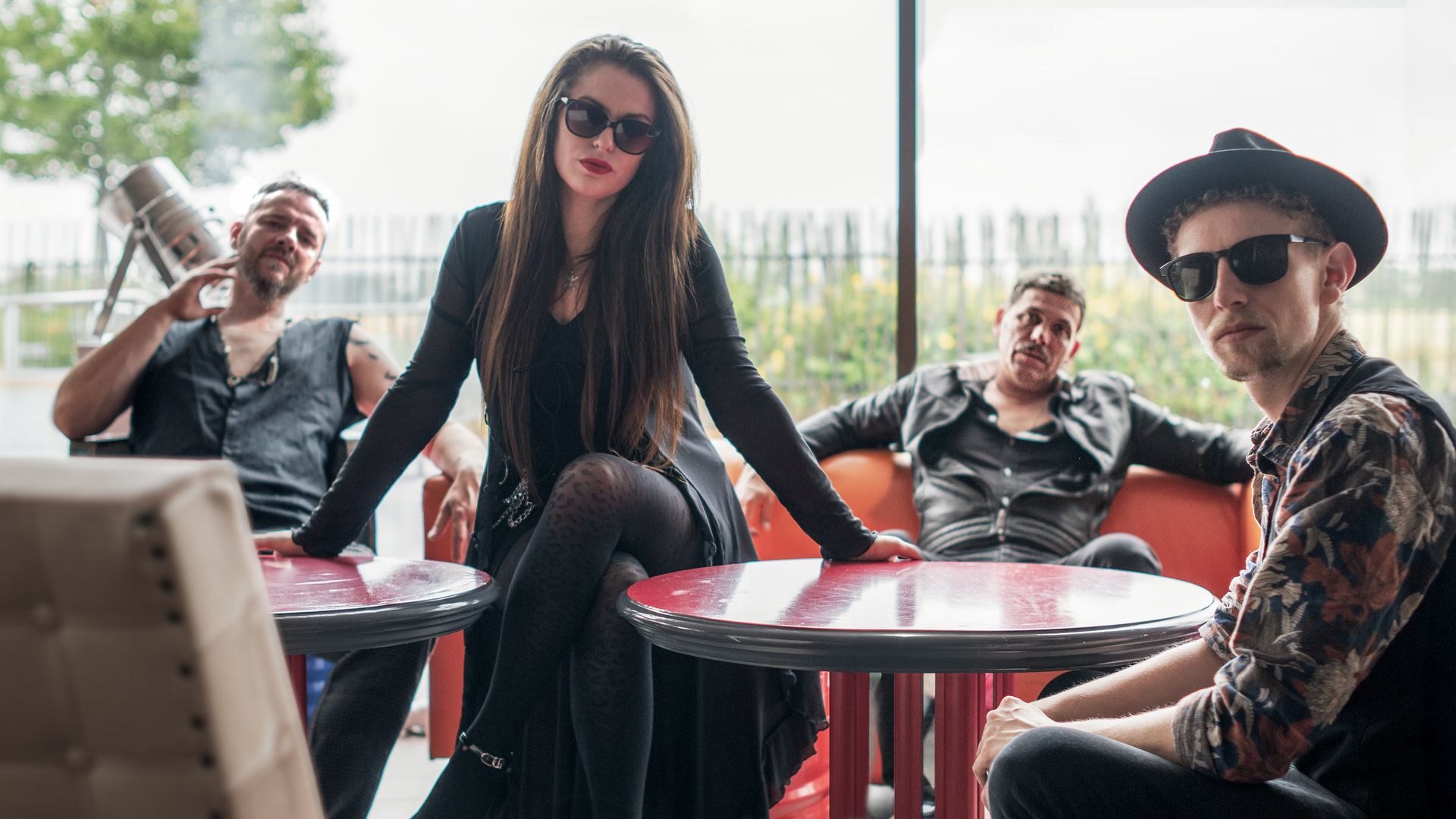 One woman wearing sunglasses sitting between two reead tables with with two male band mates in letter sits behind and another bandmate wearing hat an gasses sits on the left side of woman facing away