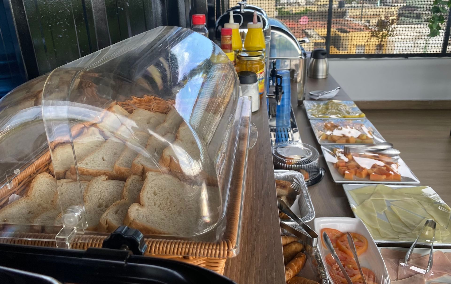 A buffet with bread under a plastic cover and various food items on plates.