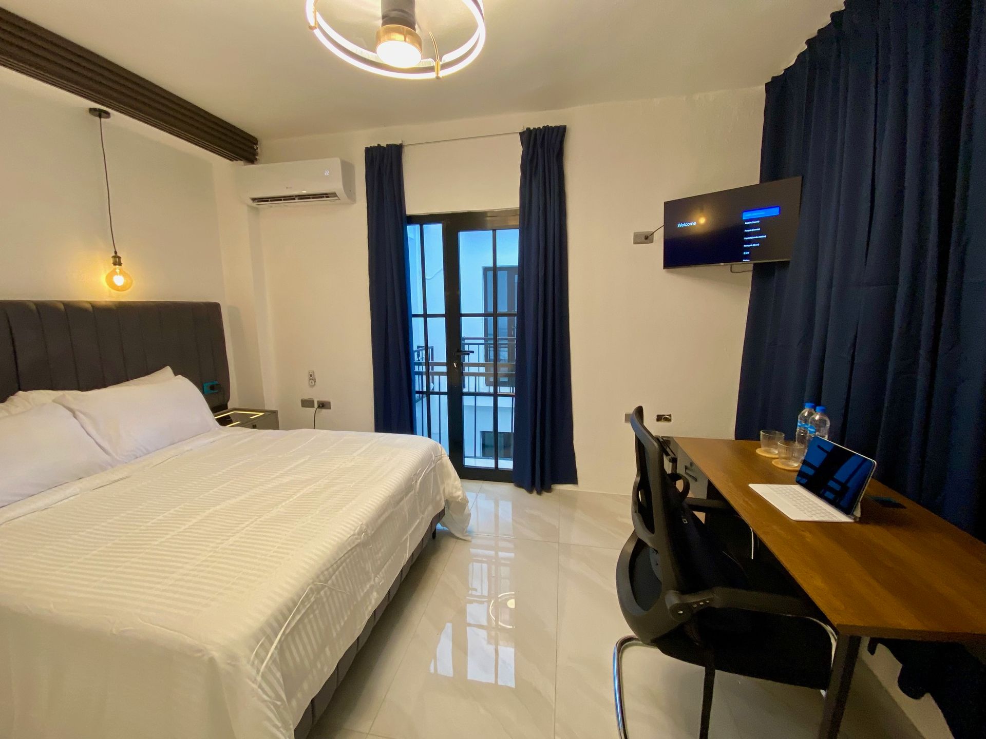 Hotel room with a bed, desk, TV, and balcony access. Dark blue curtains and white walls.
