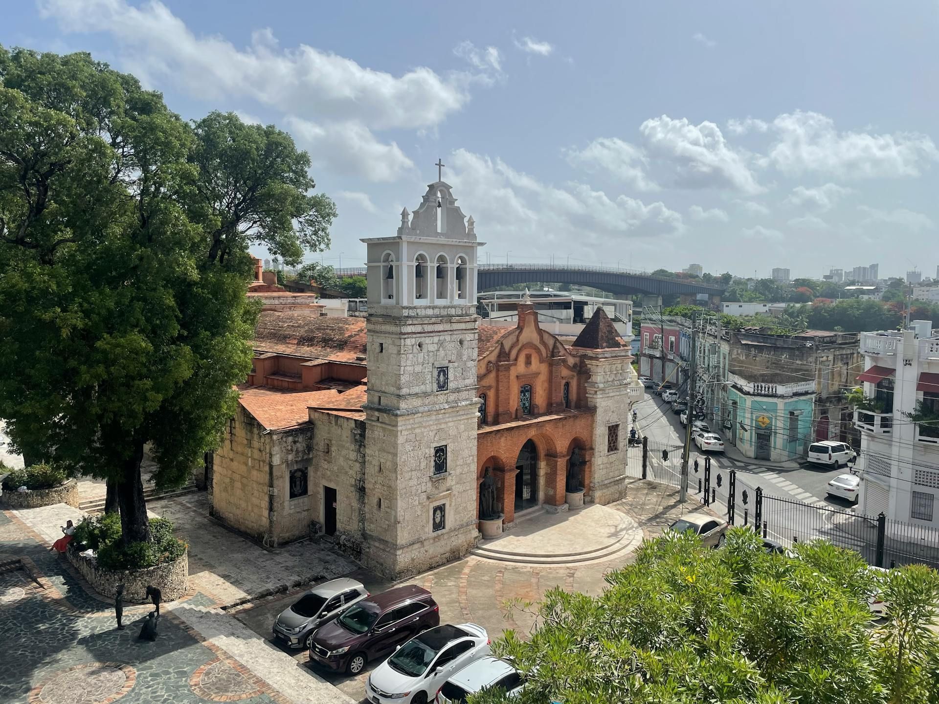 Stone church with bell tower and red-tiled roof in city square, cars parked in front, trees and buildings around.