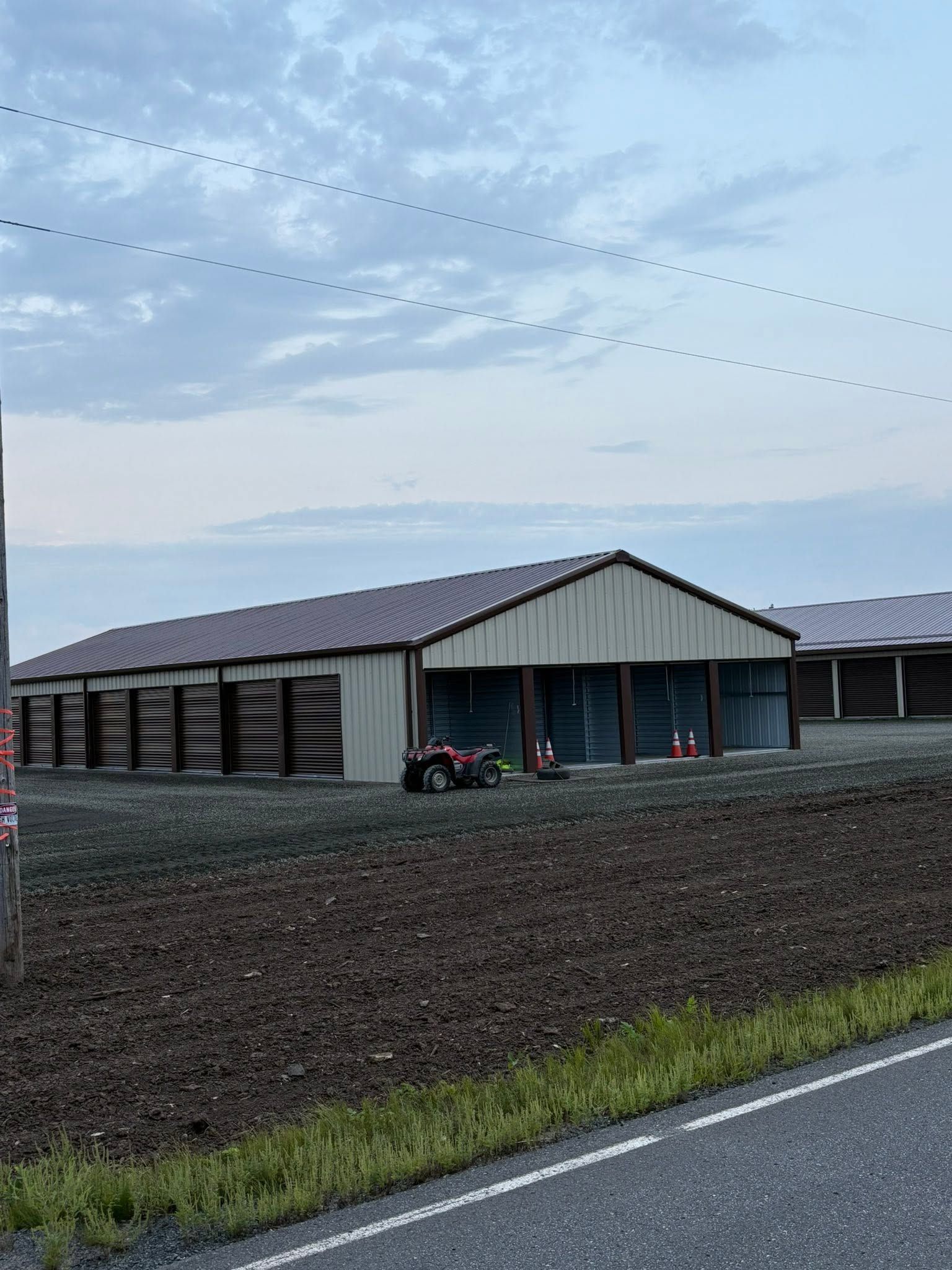 Rural building with a red tractor beside it, under a cloudy sky near a road and plowed field
