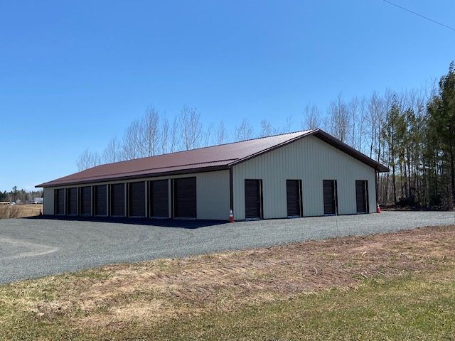 Long gray barn-like building with a red roof on a gravel lot under a blue sky