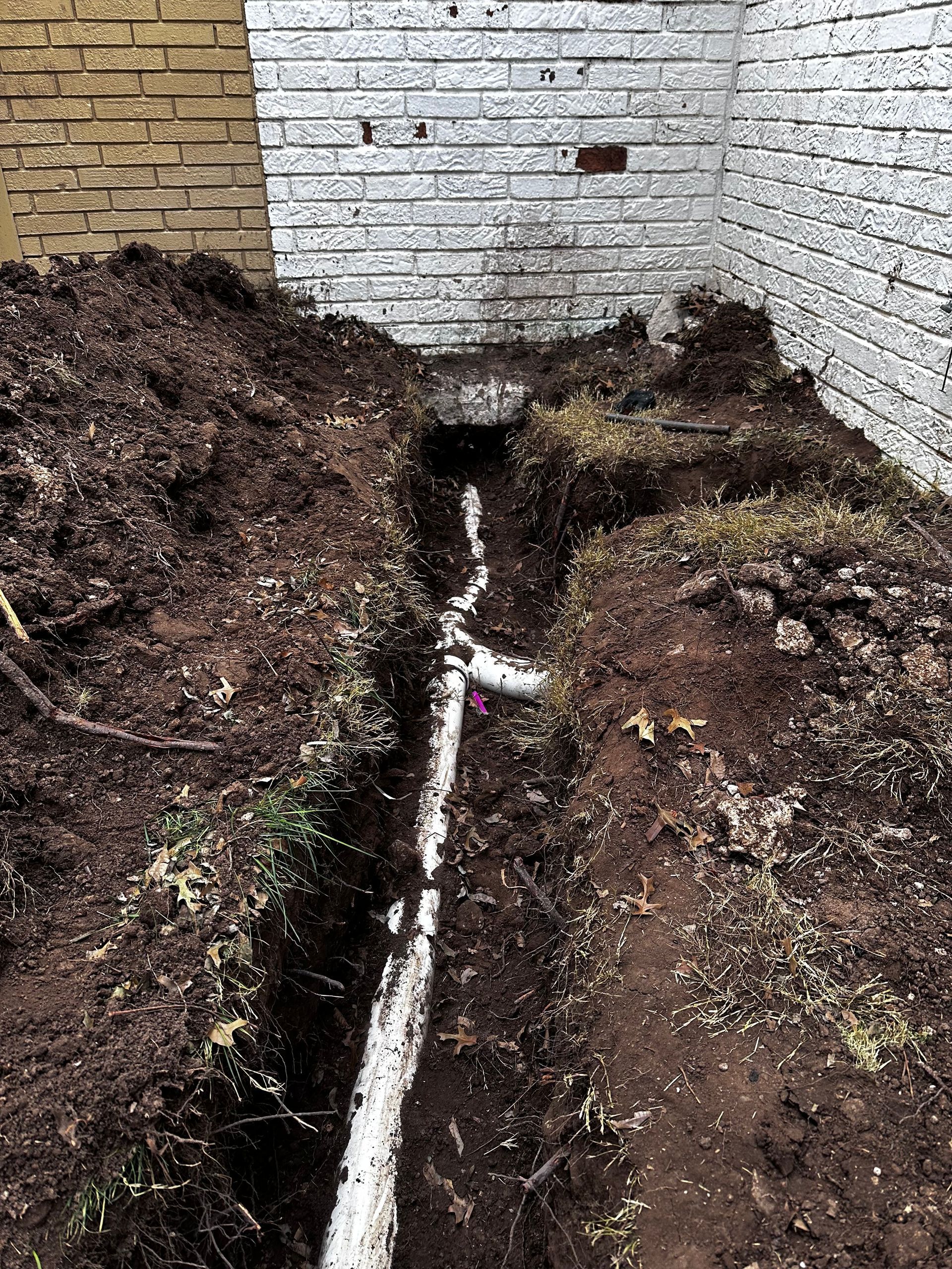 A drain pipe is being installed in the dirt in front of a brick wall.