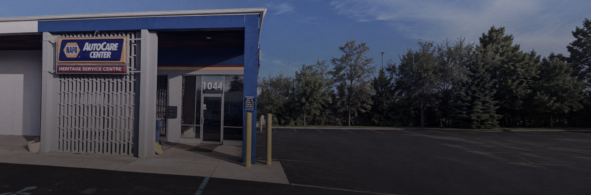 Small storefront with a sign, white fence, and empty parking lot under a cloudy sky | Heritage Service Centre