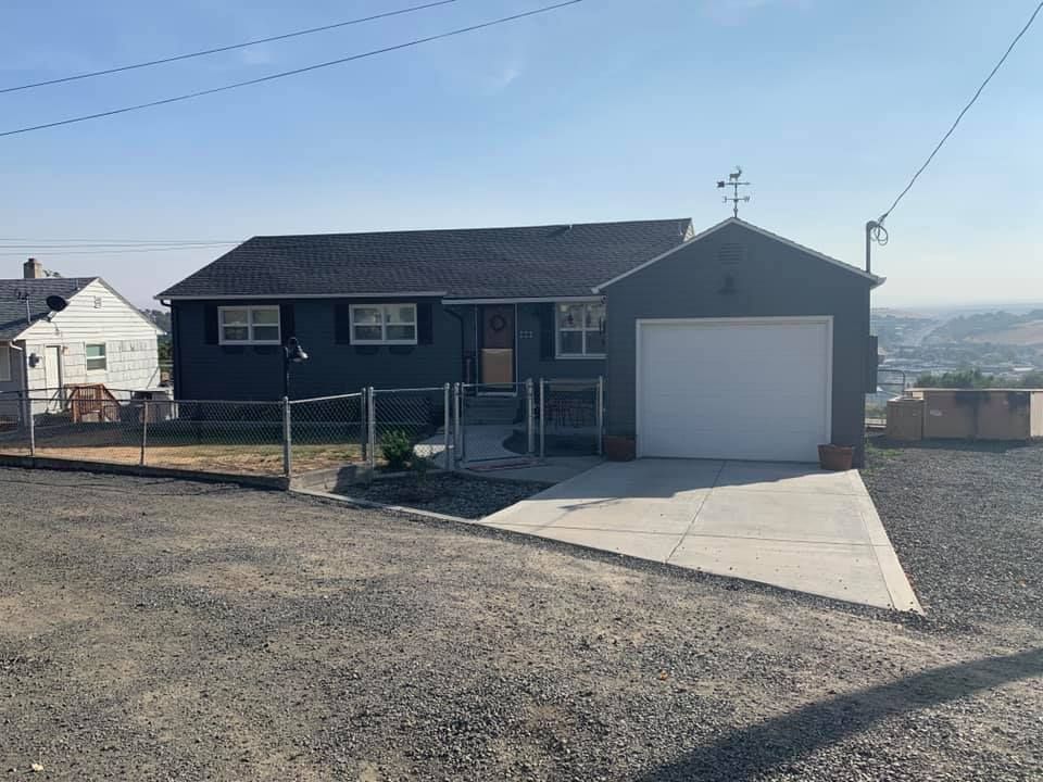 Blue house with attached garage, gravel driveway, and fence. City view in background.