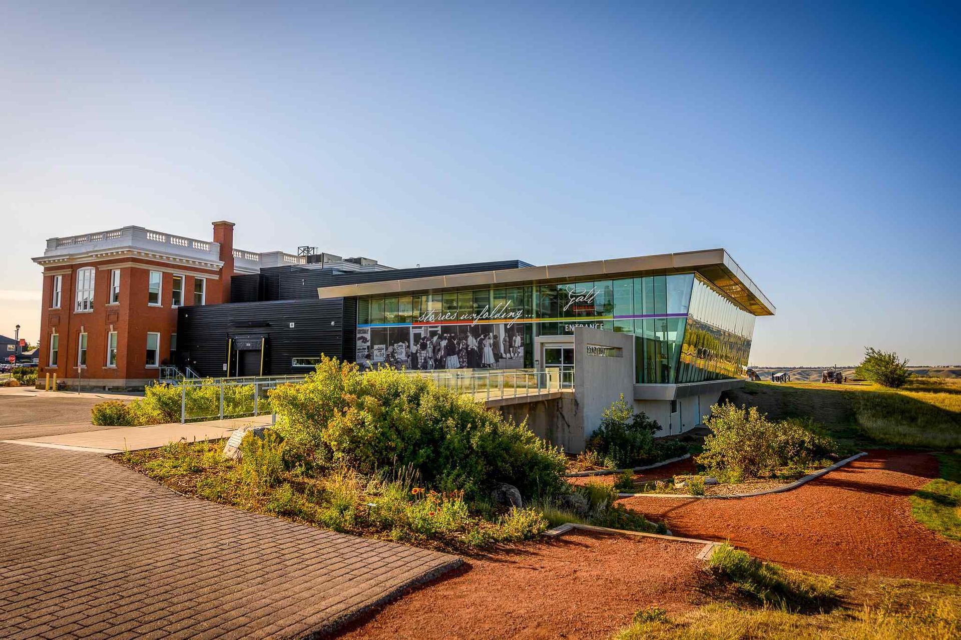 Modern glass building beside a brick structure, surrounded by landscaped gardens and pathways under a clear sky