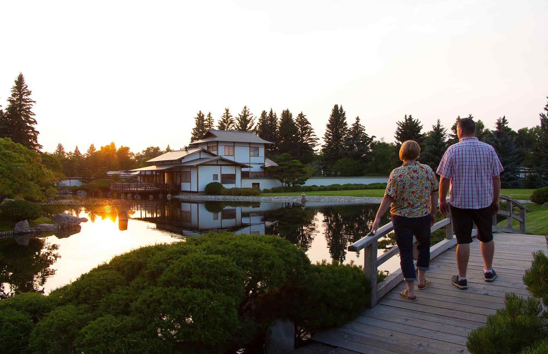 Two people walking on a boardwalk by a pond at sunset, with a house and trees reflected in the water.