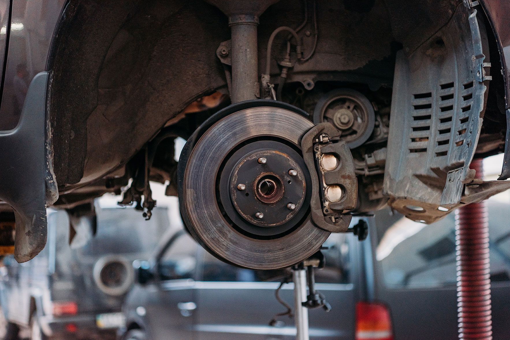 Car's front wheel brake system exposed, showing rotor, caliper, and suspension components, in a garage.