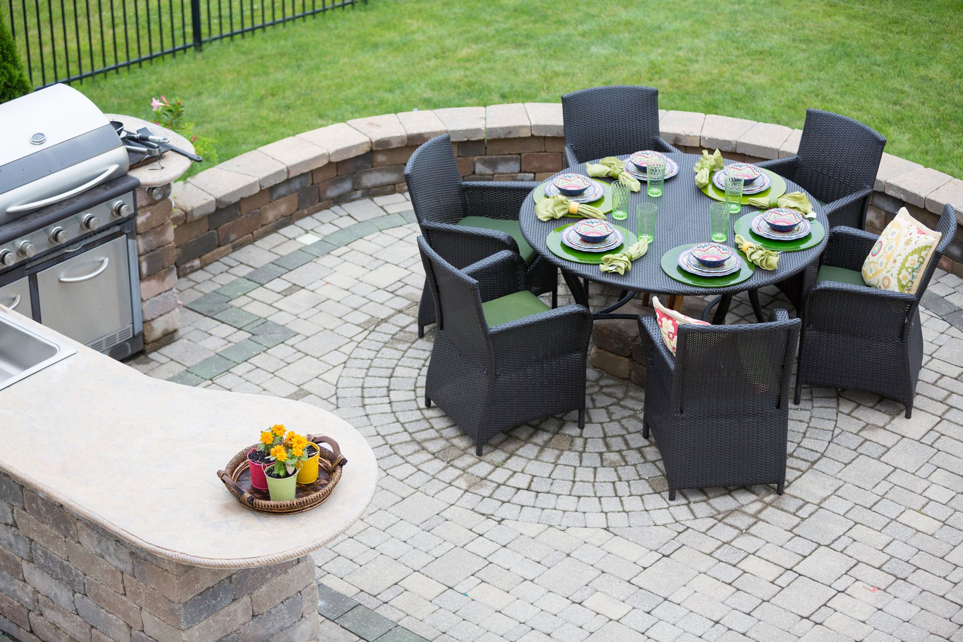 Outdoor patio with a circular table, six black wicker chairs, and a stone grill area with a counter in a grassy yard.