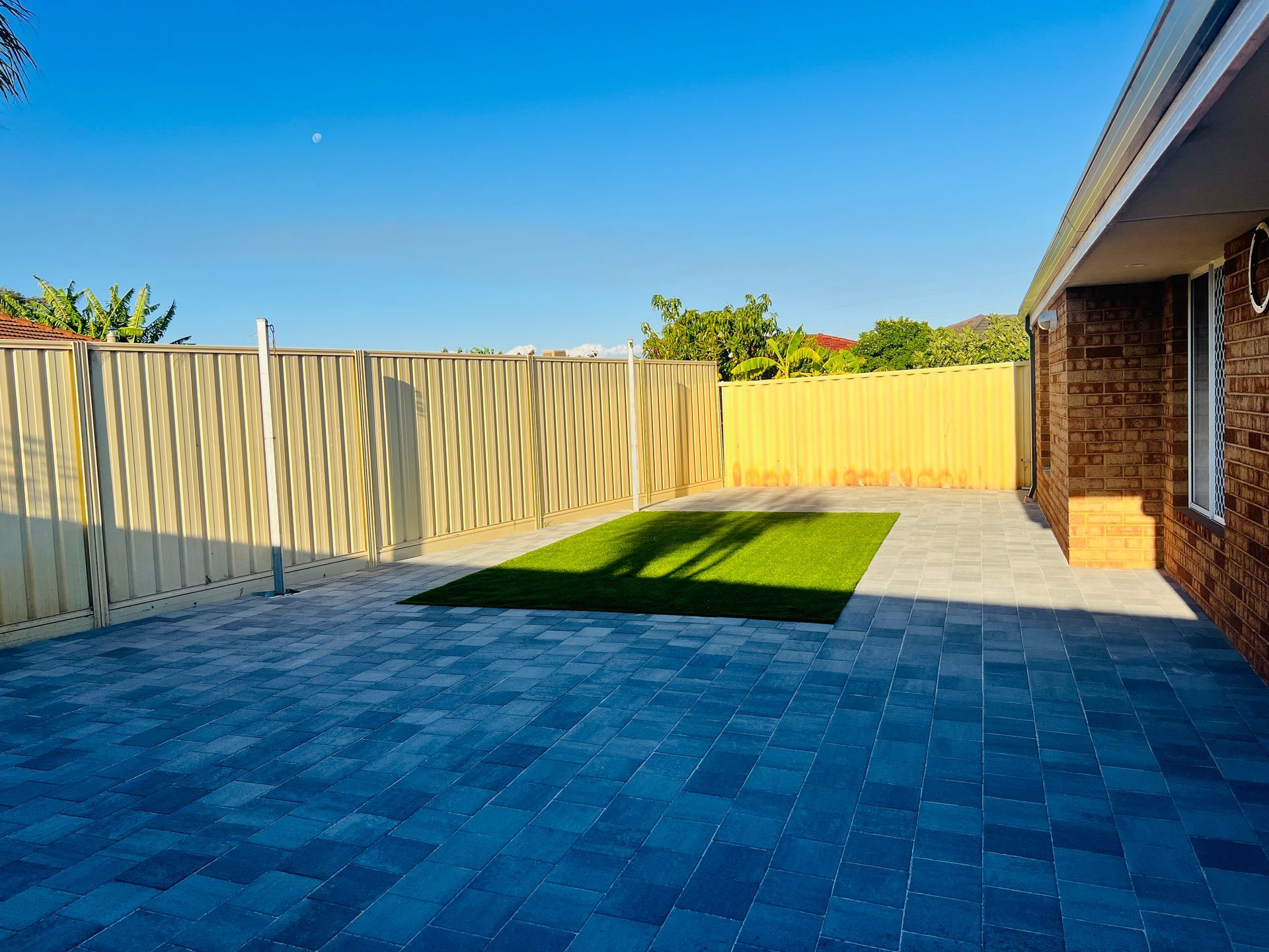 Backyard patio with blue pavers, small grass patch, tan fence, and brick house wall under a clear sky
