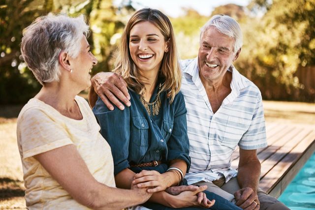 Three smiling people sit together by a pool on a sunny wooden deck.