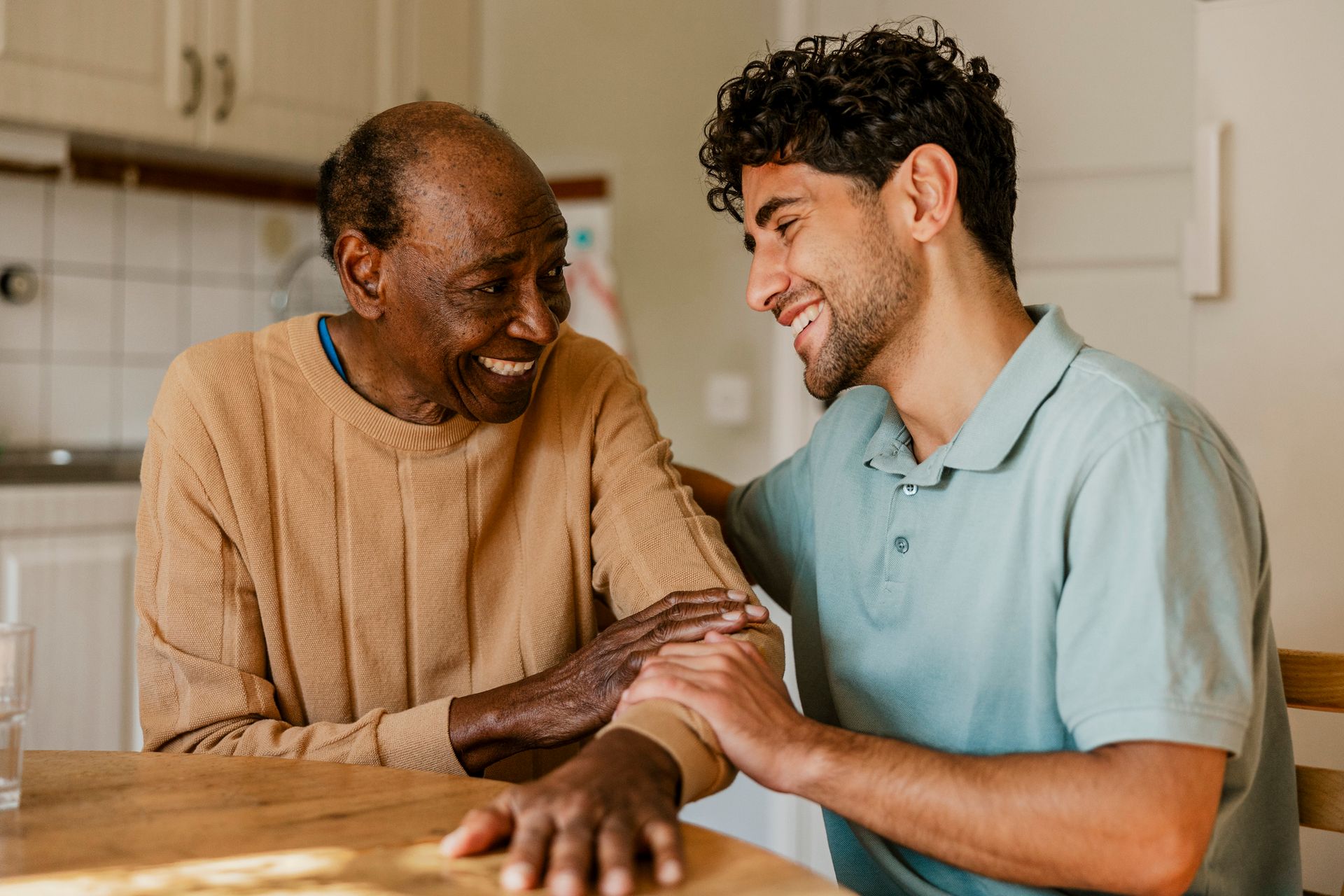 An older Black man and younger man smile, with the younger man's hand on the older man's arm, indoors. An older Black man and younger man smile, with the younger man's hand on the older man's arm, indoors.