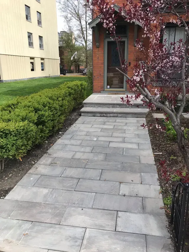 Brick walkway leading to a red brick building with a door, flanked by greenery and a small tree.