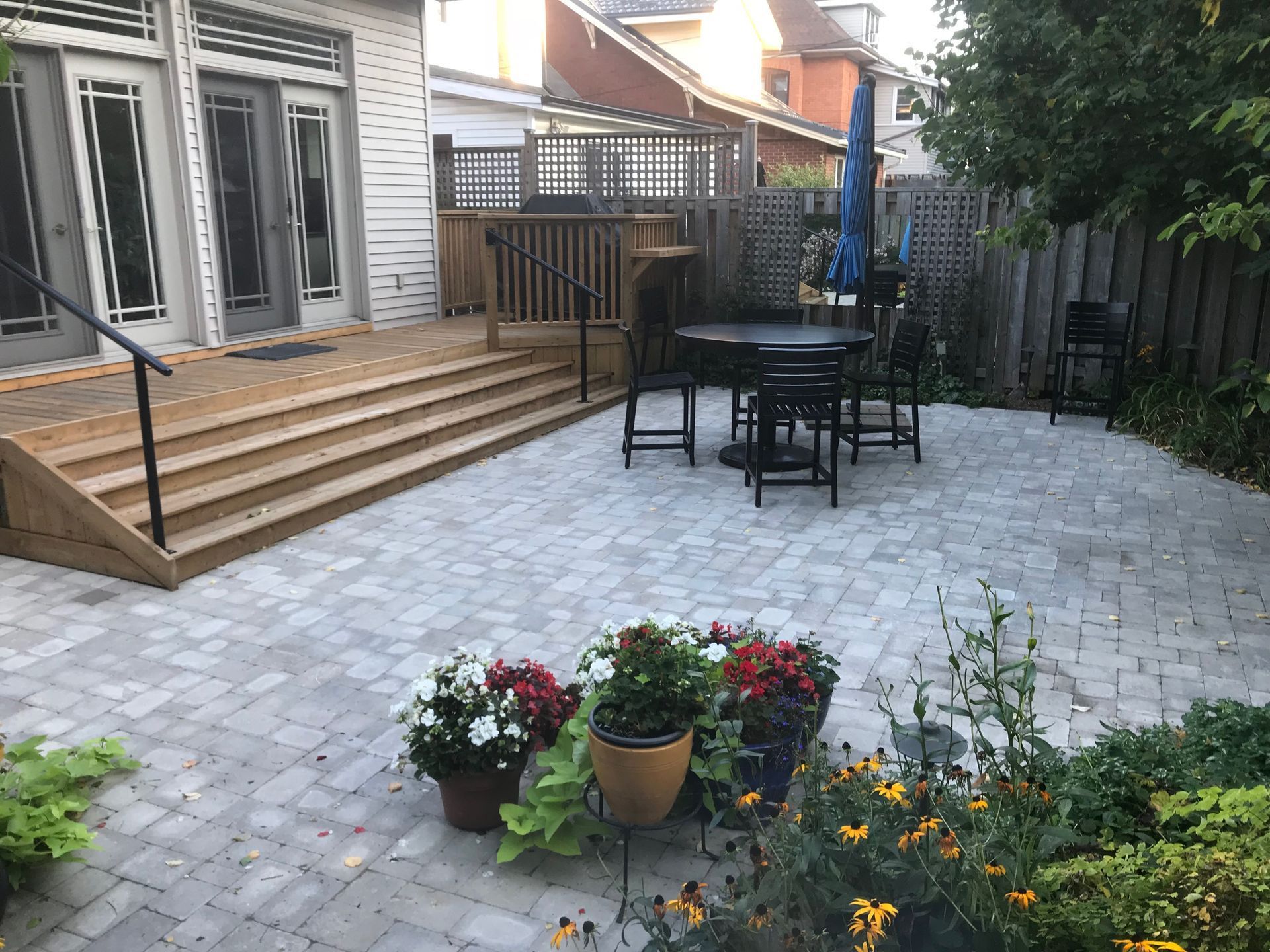 Patio with paved stones, wooden deck, table, chairs, potted flowers, and garden foliage.