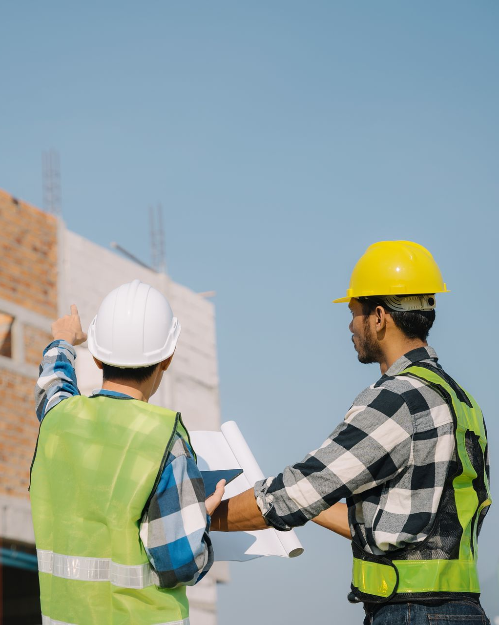 Two construction workers reviewing blueprints at a building site; one points upwards.