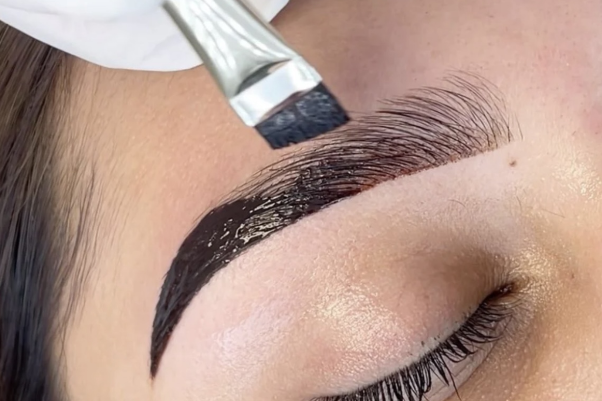 Close-up of a hand applying dark eyebrow makeup with a brush to a closed eye