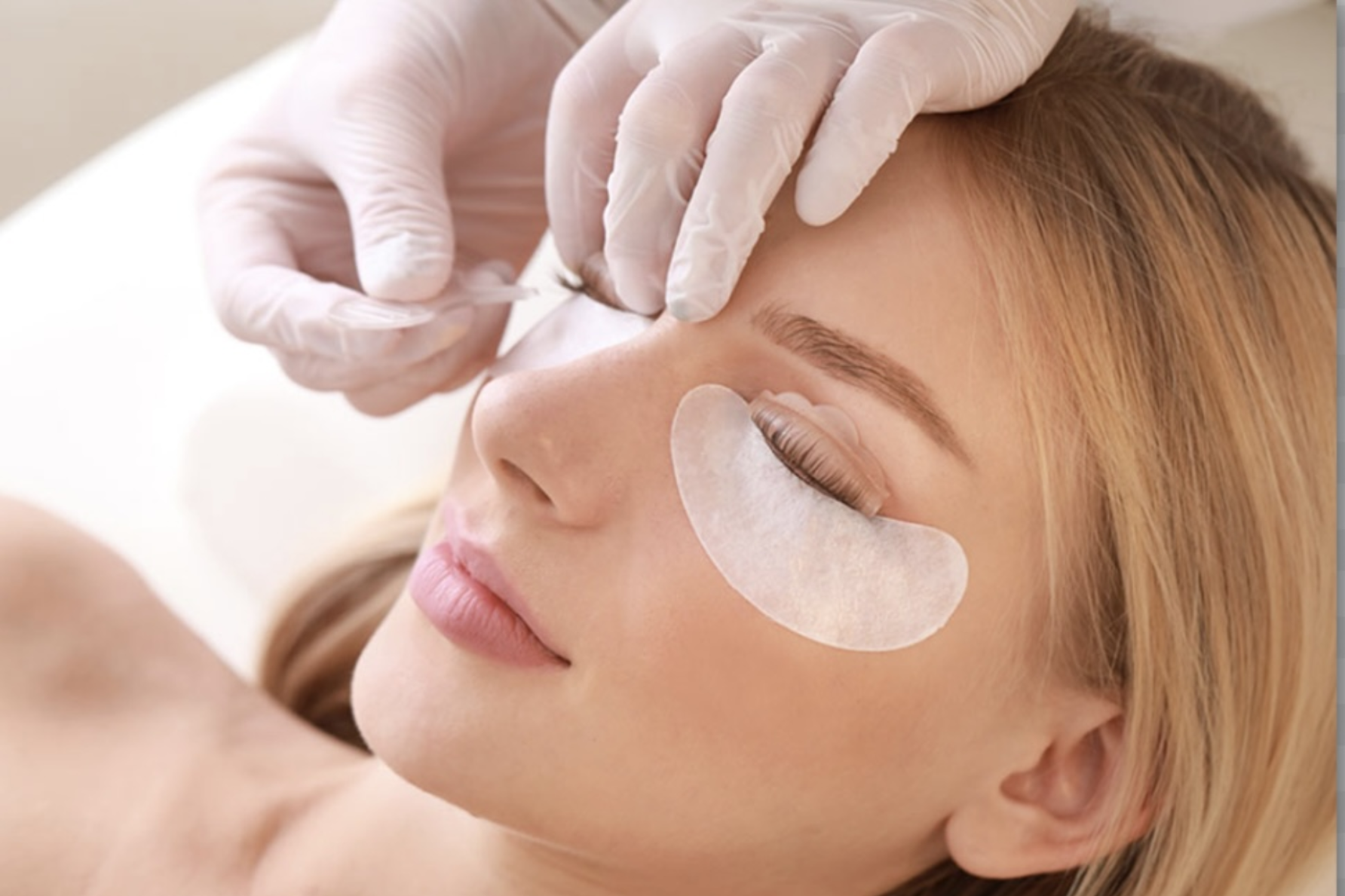 Beauty treatment with under-eye patches as gloved hands apply brow grooming to a reclining client