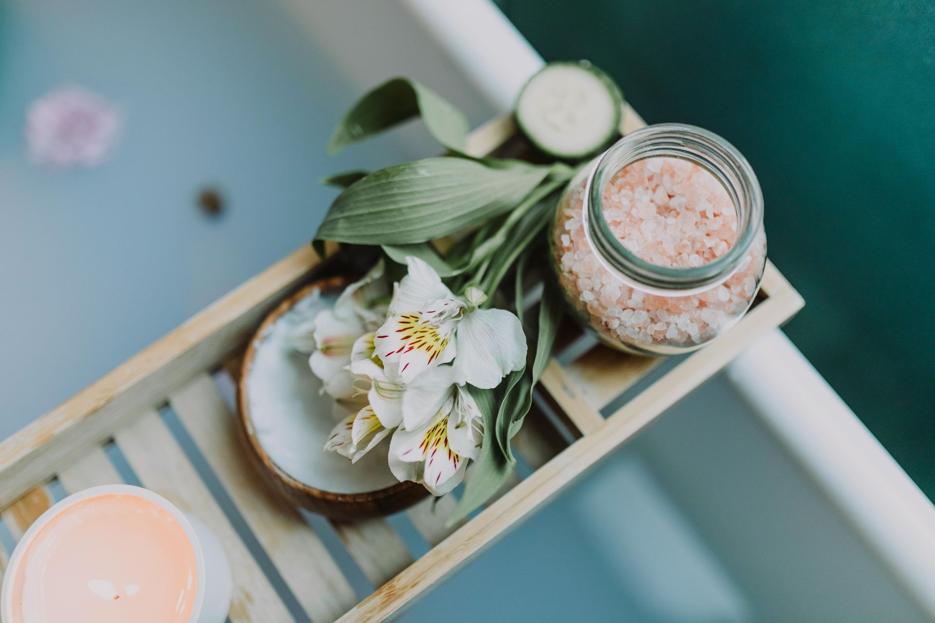 Decorative tray with flowers, leaves, cucumber slices, and a jar of pink bath salts on a blue surface