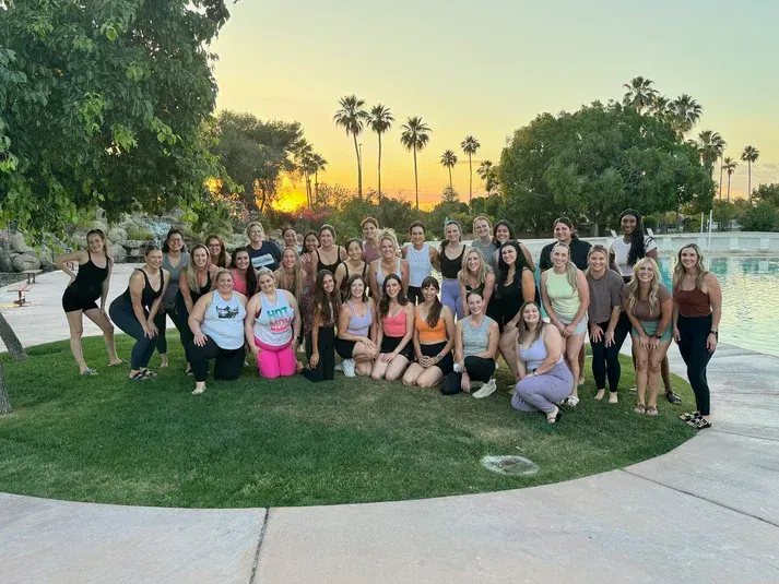 Large group posing on a grassy lawn at sunset, with palm trees and a pool in the background.