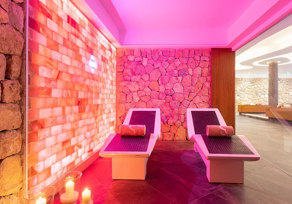 Pink-lit spa room with two massage beds, stone wall, and large windows overlooking a sunny view