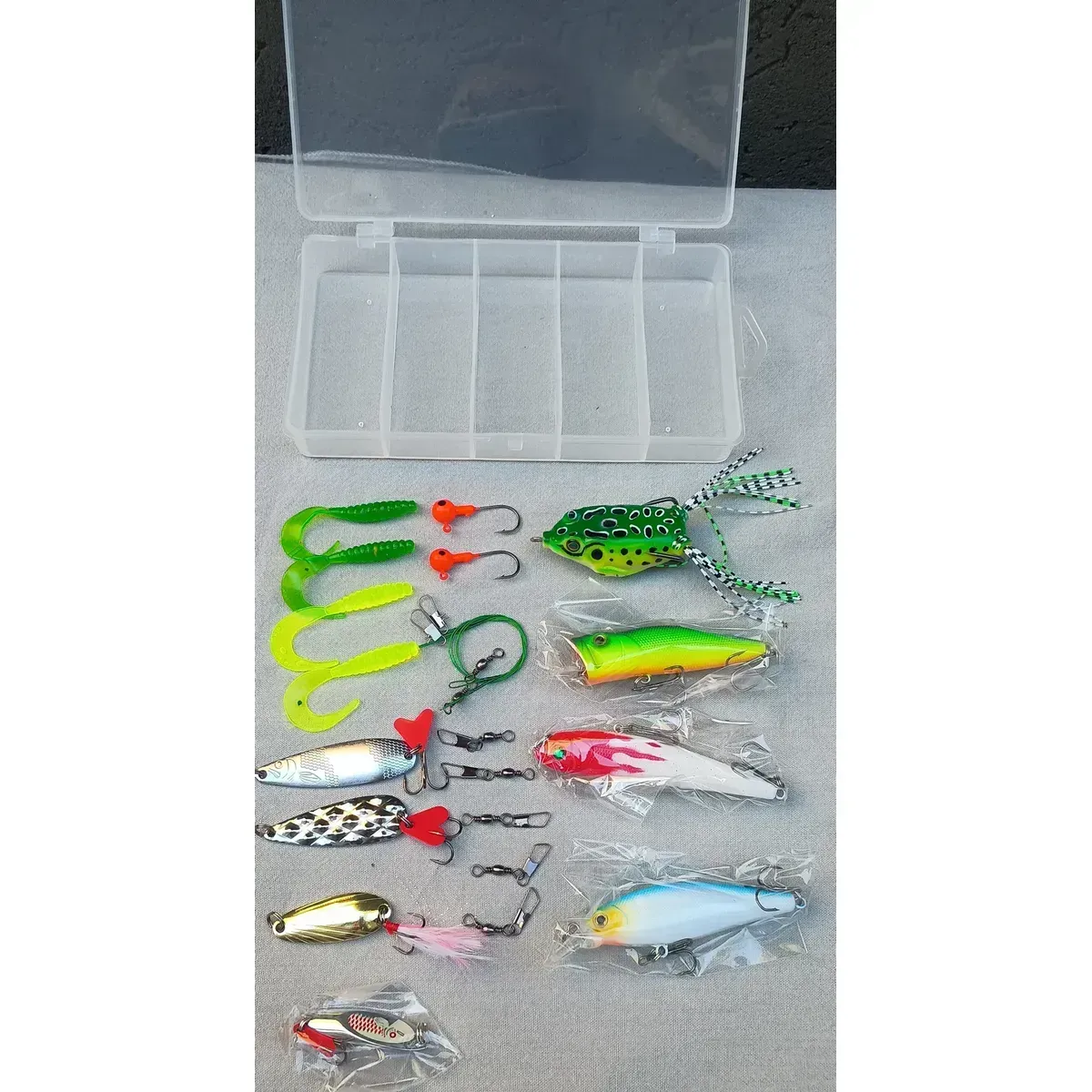 Caja de plástico para aparejos de pesca con cinco señuelos de colores sobre una mesa de madera al aire libre