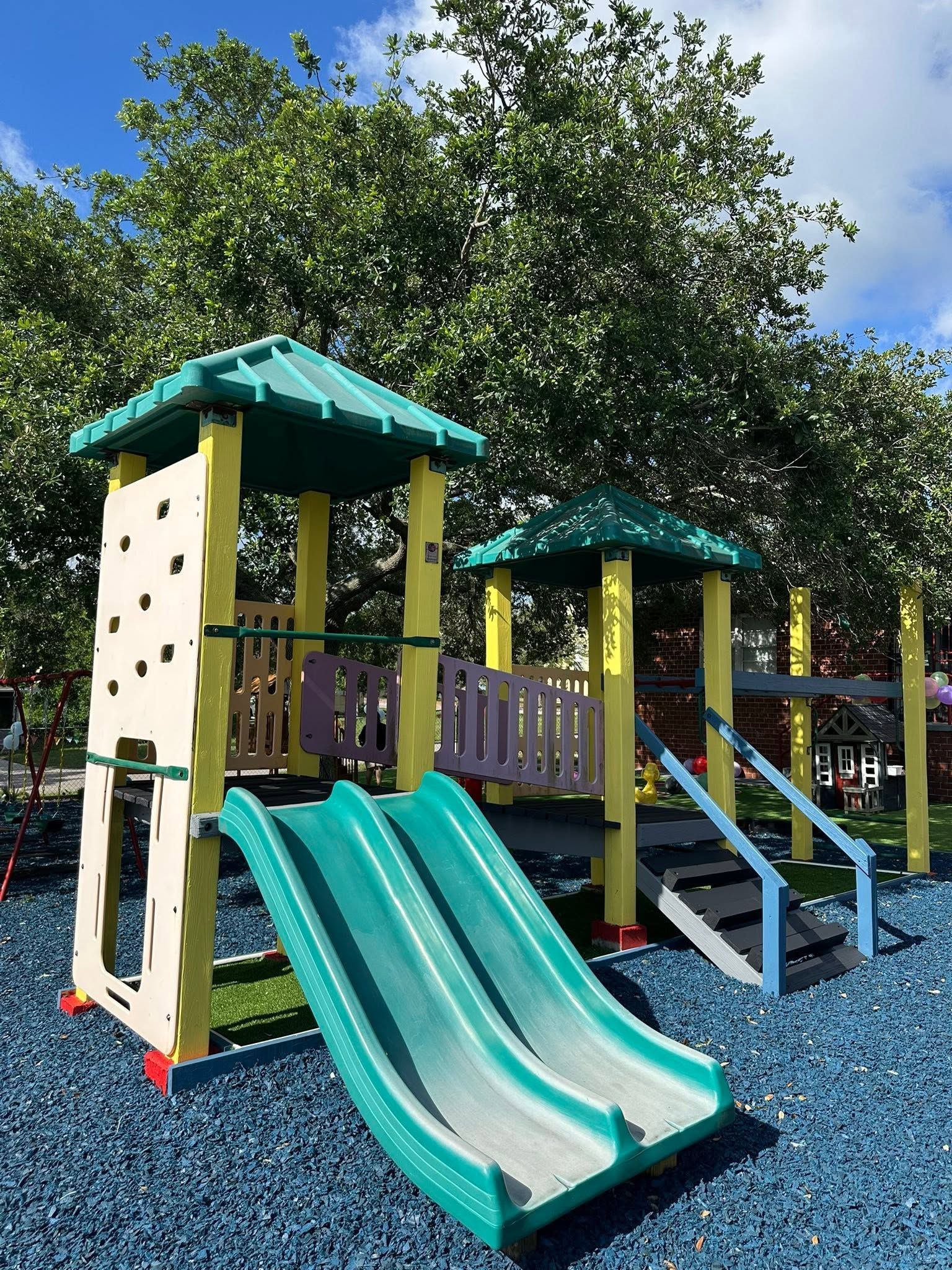 Colorful playground with two slides, climbing structure, and blue ground under trees and a sunny sky