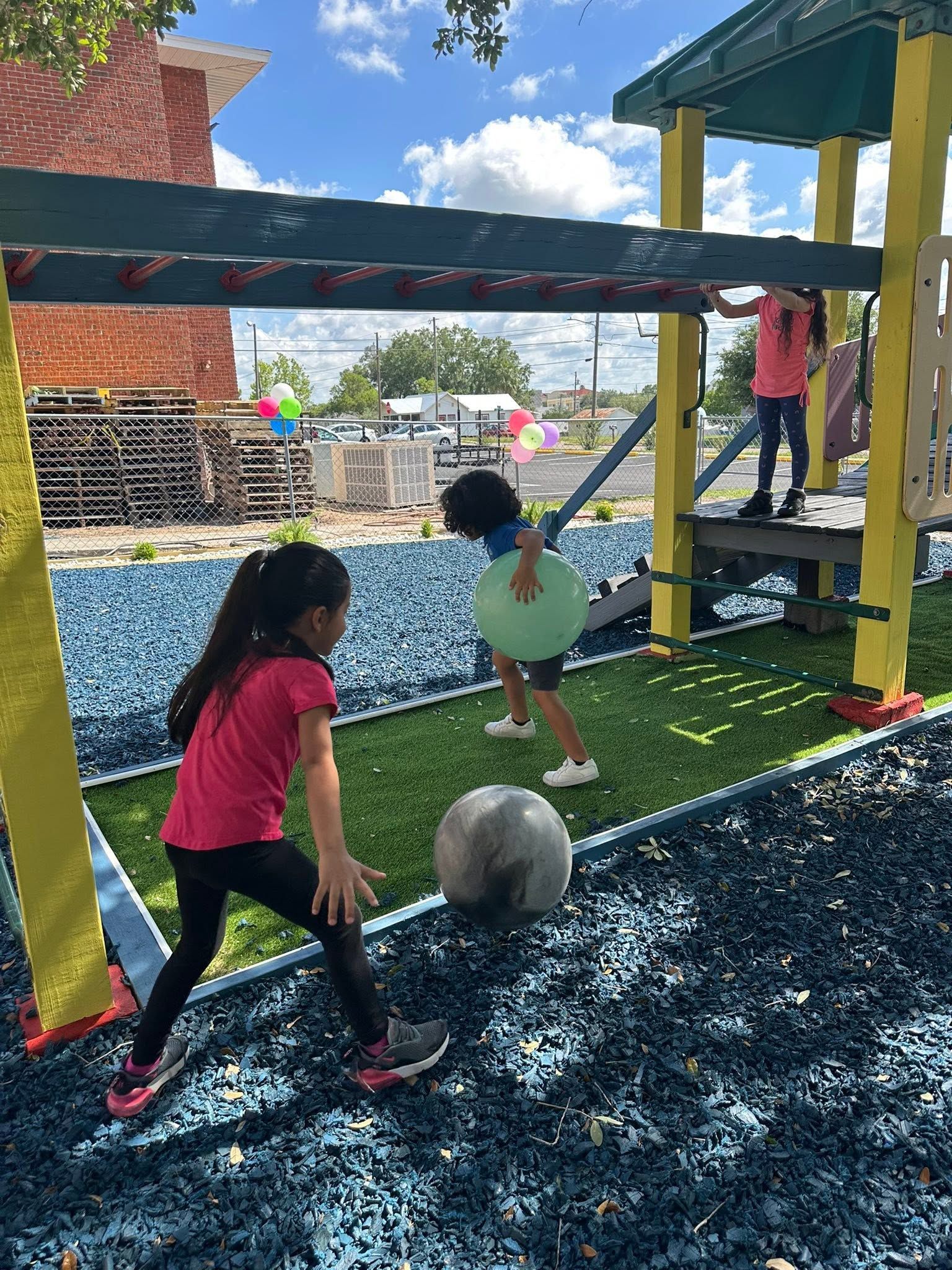 Children playing under a playground structure, with a rope swing and a large ball on rubber flooring.