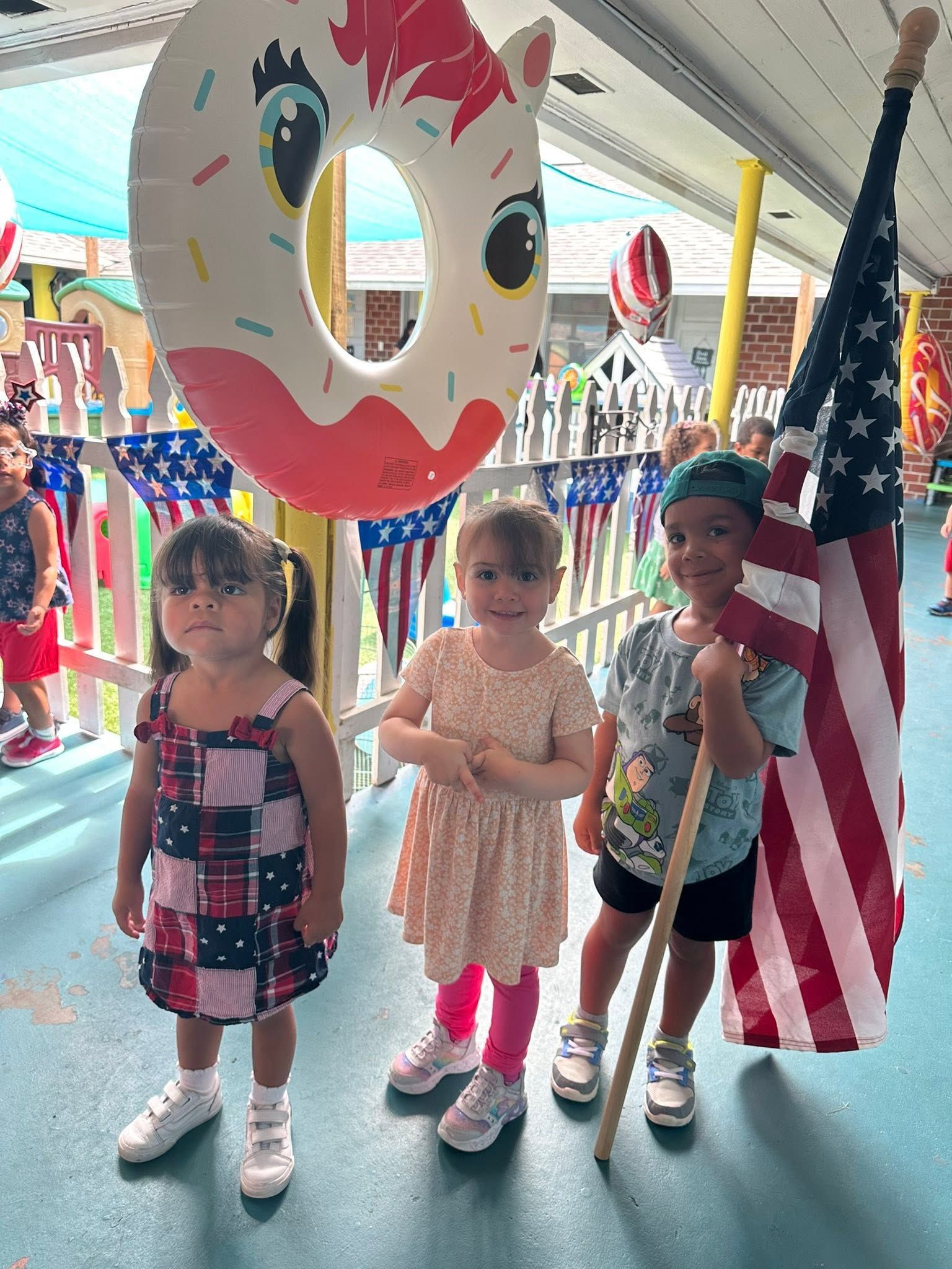 Three children pose indoors beside a large smiley face cutout and an American flag.