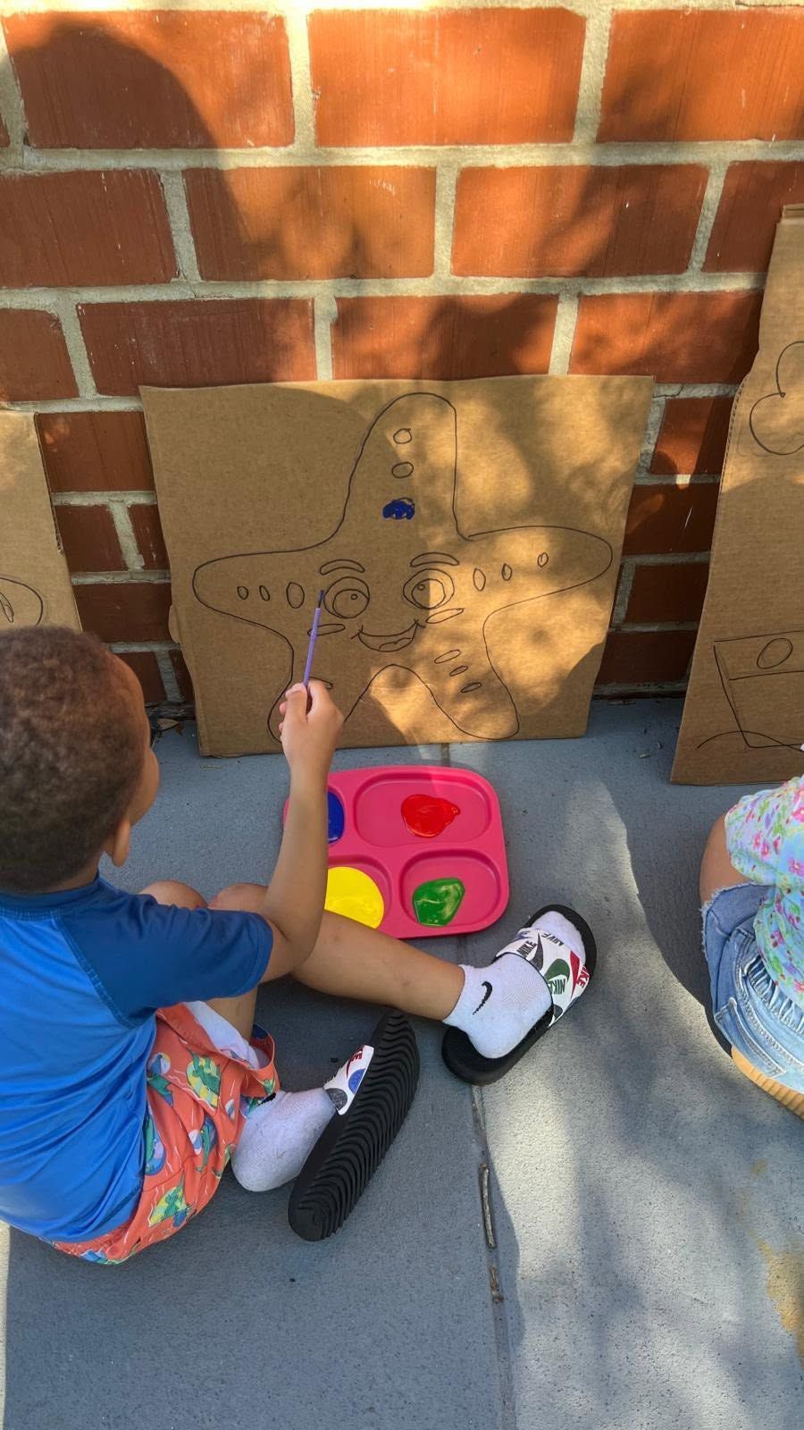 Child playing a shape-matching game on cardboard against a brick wall with colorful toy pieces nearby