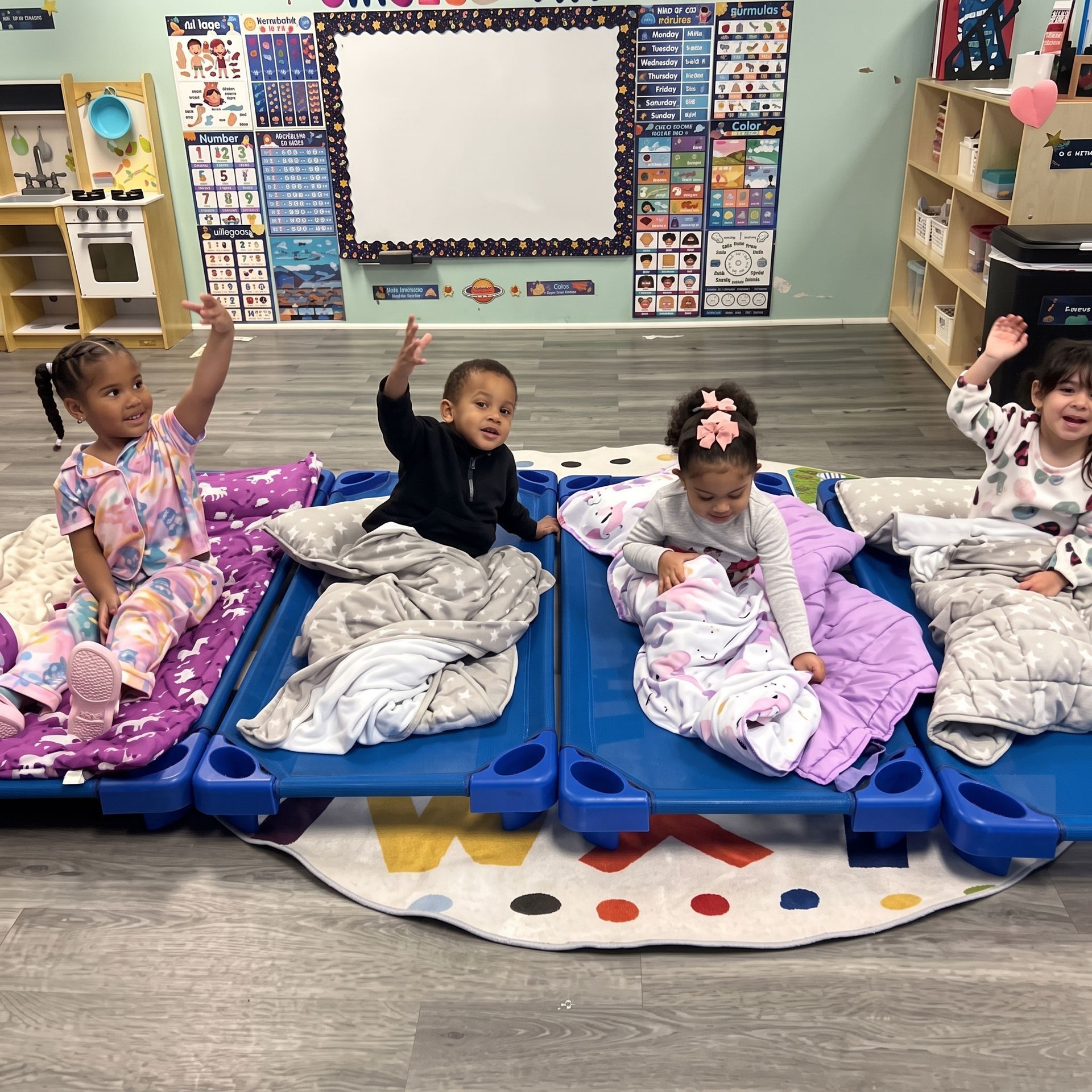 Four children lie on mats in a classroom, smiling and waving during nap time.
