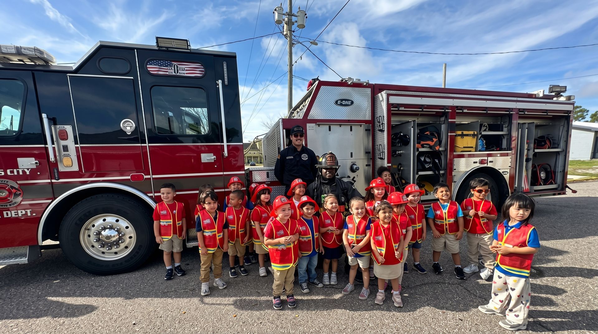 Children in red shirts pose in front of two fire trucks on a sunny day.