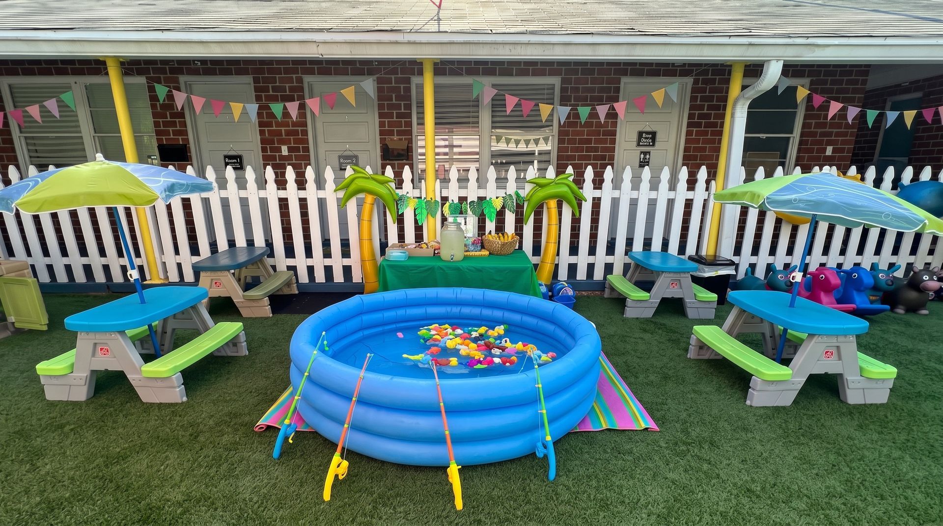 Colorful backyard play area with a blue kiddie pool, toy umbrellas, and a white picket fence