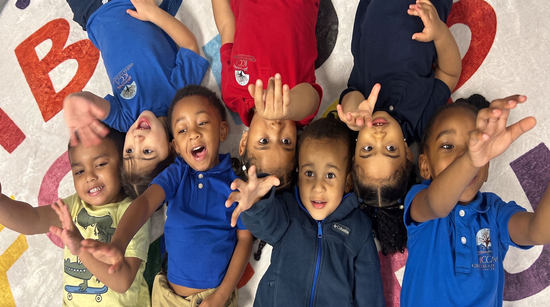 Children lying in a circle on a classroom floor, smiling and reaching toward the camera.