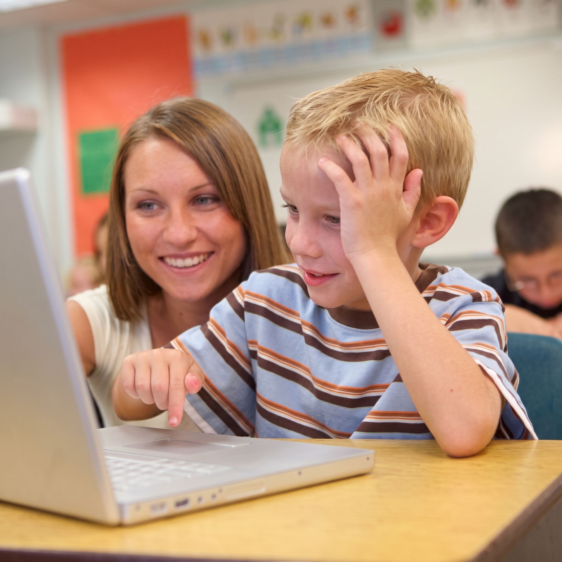 Woman and boy using a laptop in a classroom, smiling and pointing at the screen.
