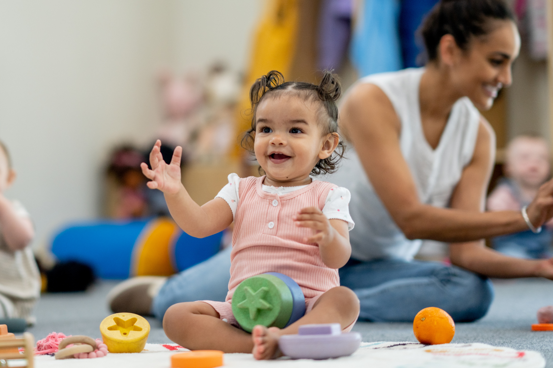 Smiling baby in pink romper playing with colorful toys on a play mat, with adults in the background