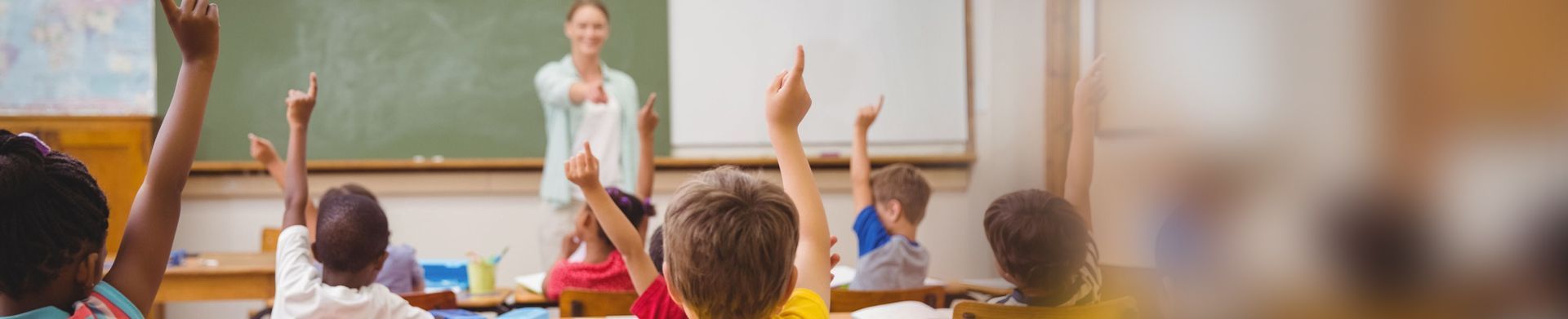 Teacher standing at front of classroom with students raising hands in a bright room