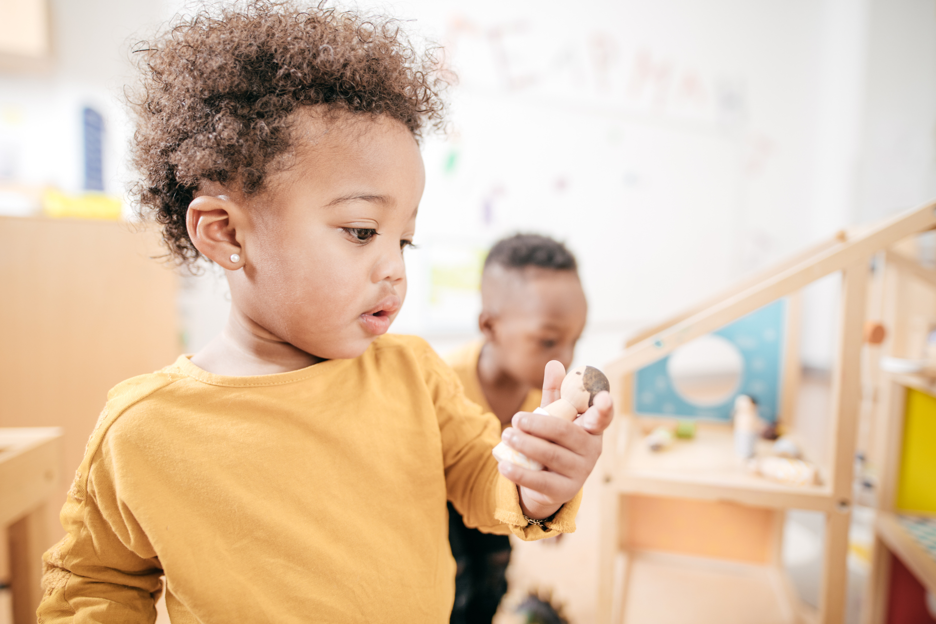 Child in a mustard shirt looking at a small object in a bright classroom, with another child blurred in the background.
