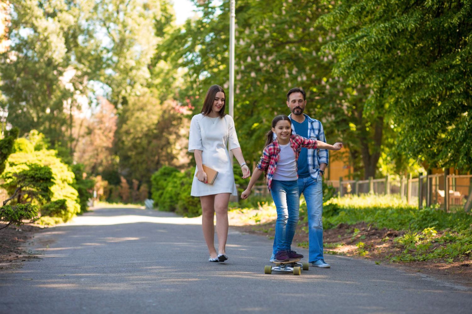 Two adults and a child riding a skateboard on a tree-lined road