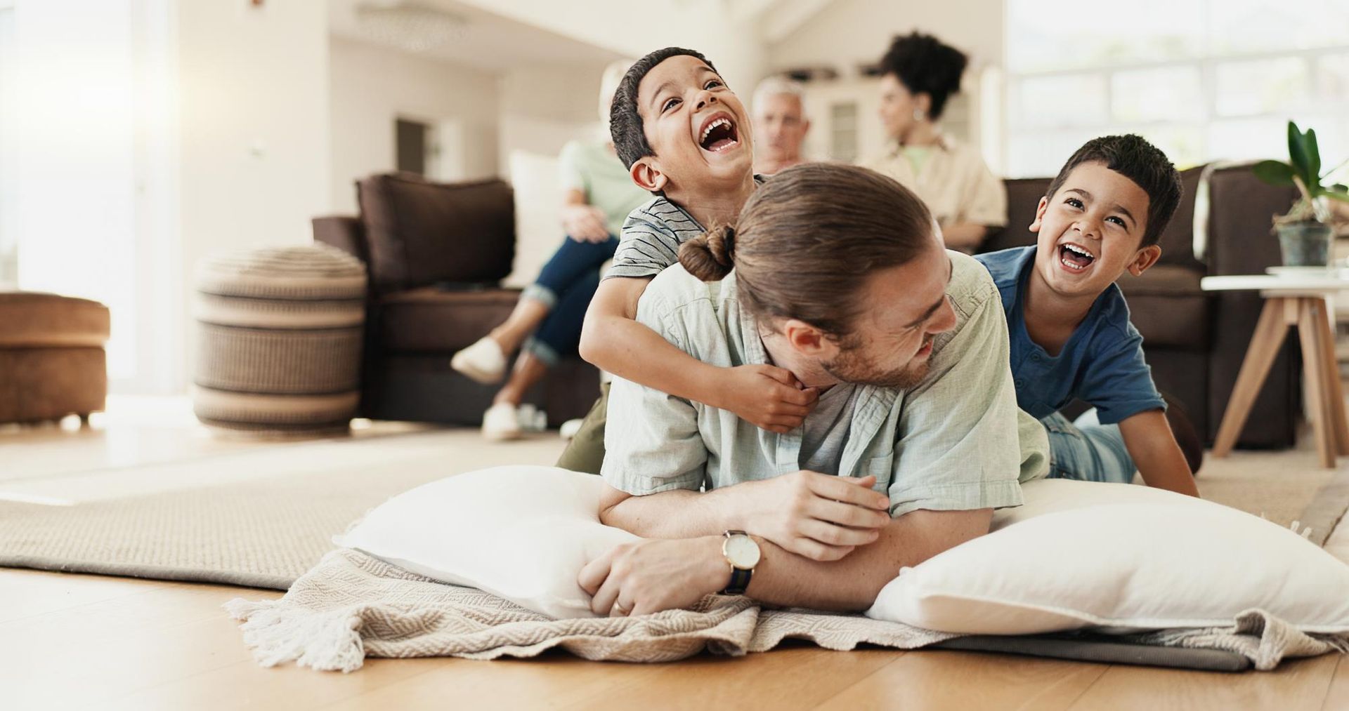 Three smiling children playfully wrestling on a living room floor with pillows.